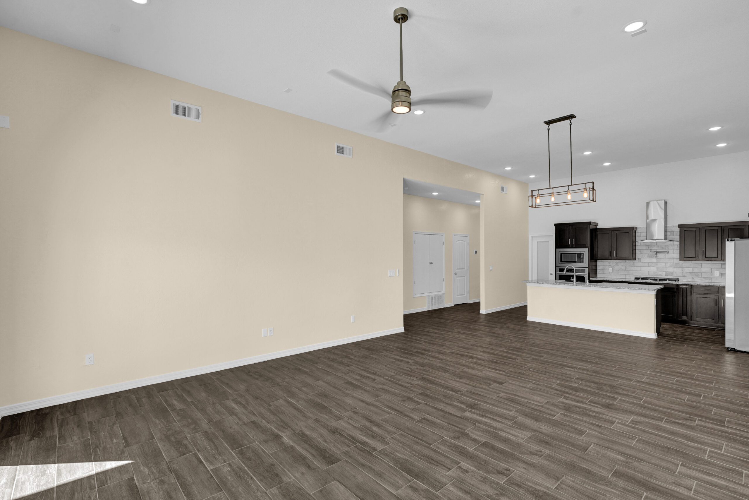 Open-concept living and kitchen space in The Lavender model by Zia Homes, featuring wood-look tile flooring, dark cabinetry, and modern ceiling fixtures.