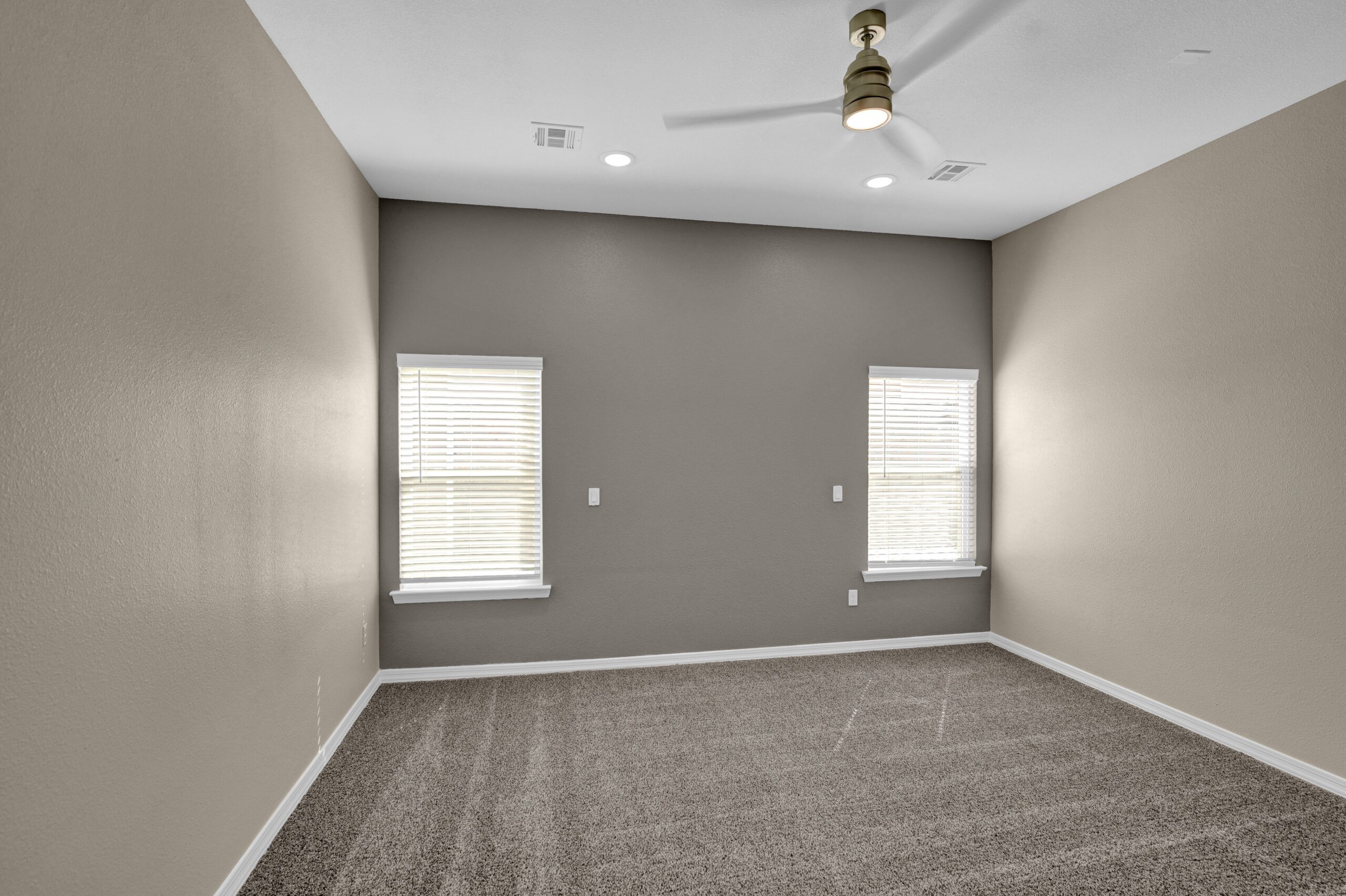 Bedroom in The Lavender model by Zia Homes featuring a modern ceiling fan, two tall windows with blinds, and plush carpeting against a neutral accent wall.