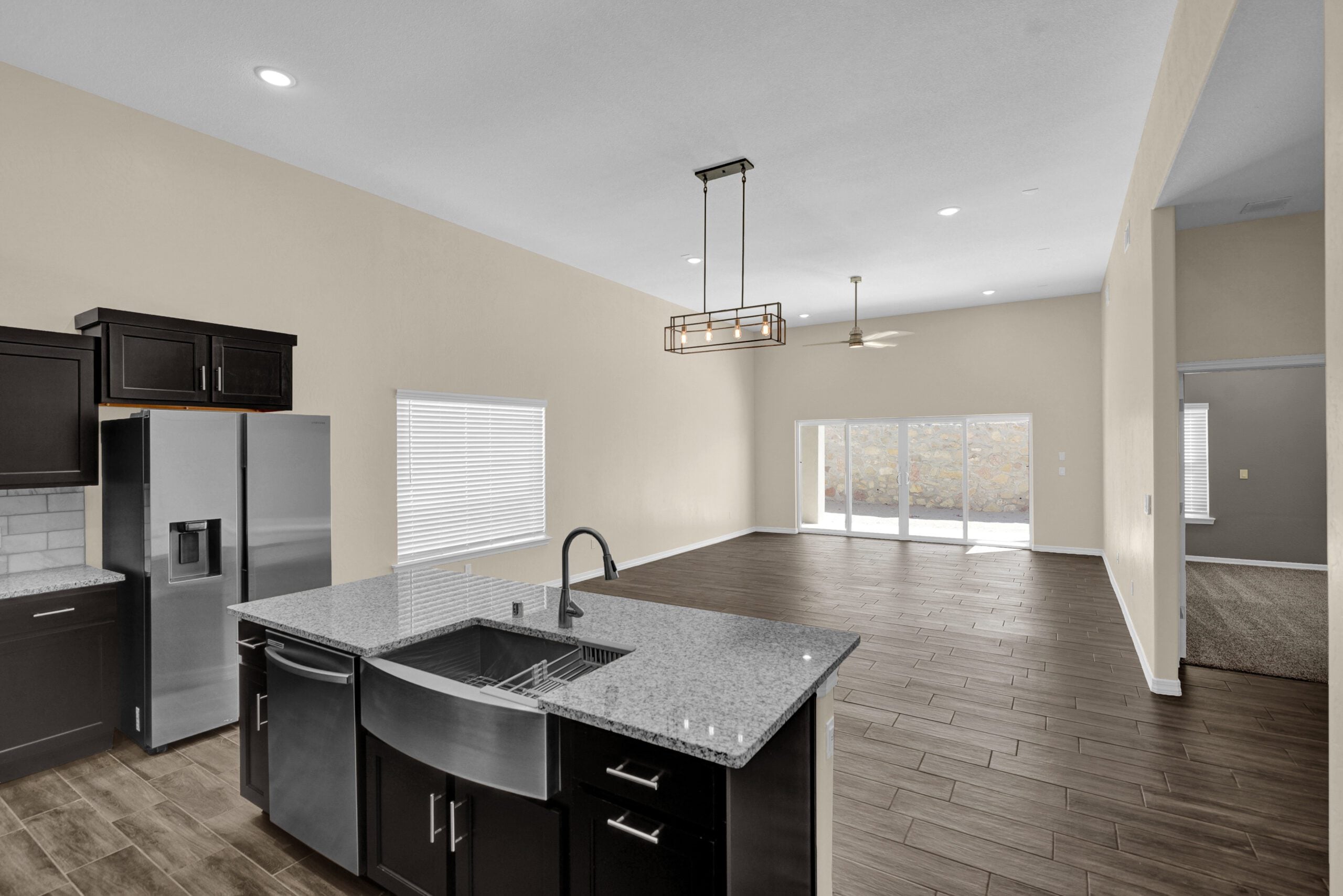 Wide-angle view from The Lavender model kitchen showing a granite island with farmhouse sink, open living area with wood-look tile flooring, and large sliding glass doors.