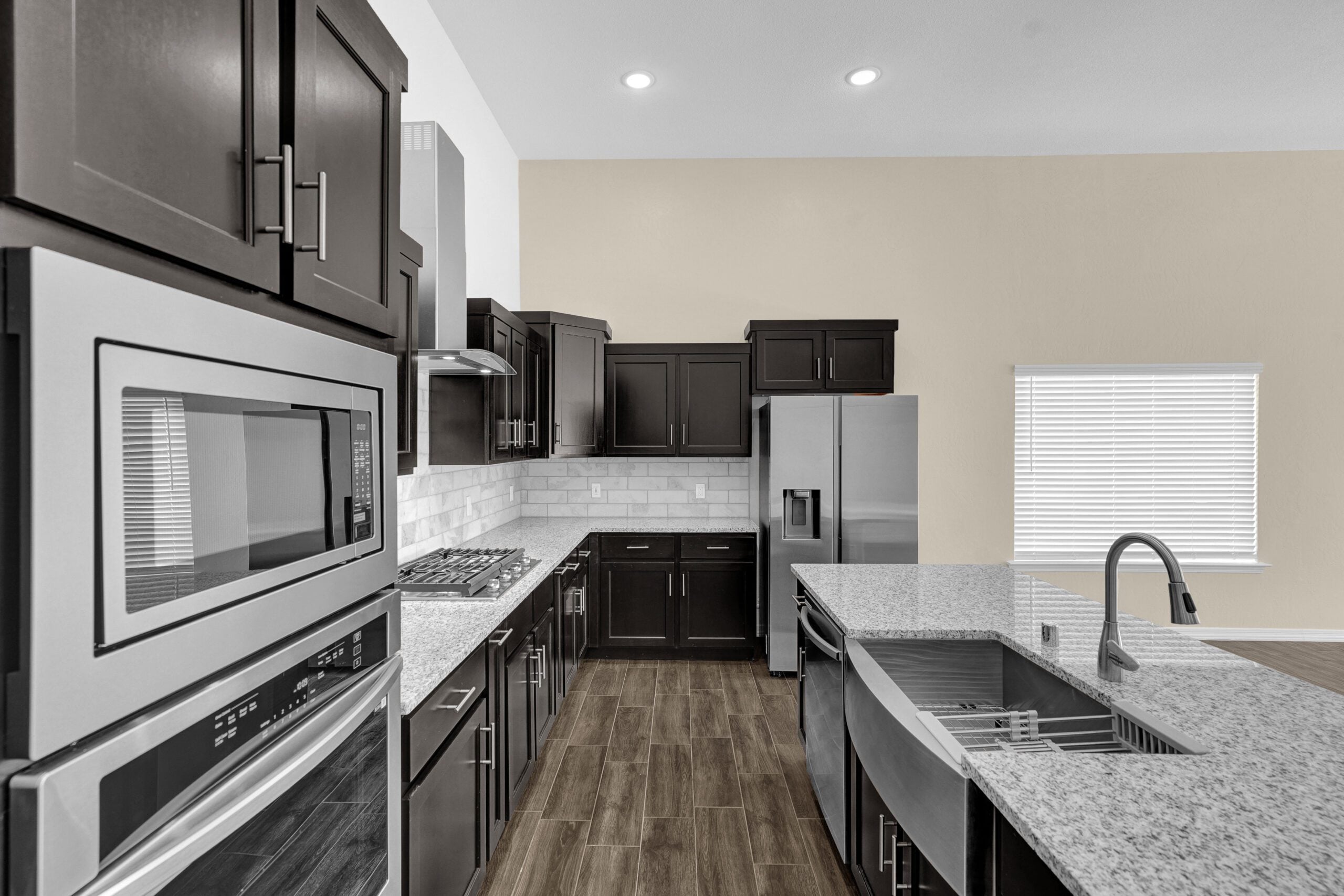 Close-up view of The Lavender kitchen by Zia Homes featuring a granite island with farmhouse sink, stainless steel appliances, and dark cabinetry with subway tile backsplash.