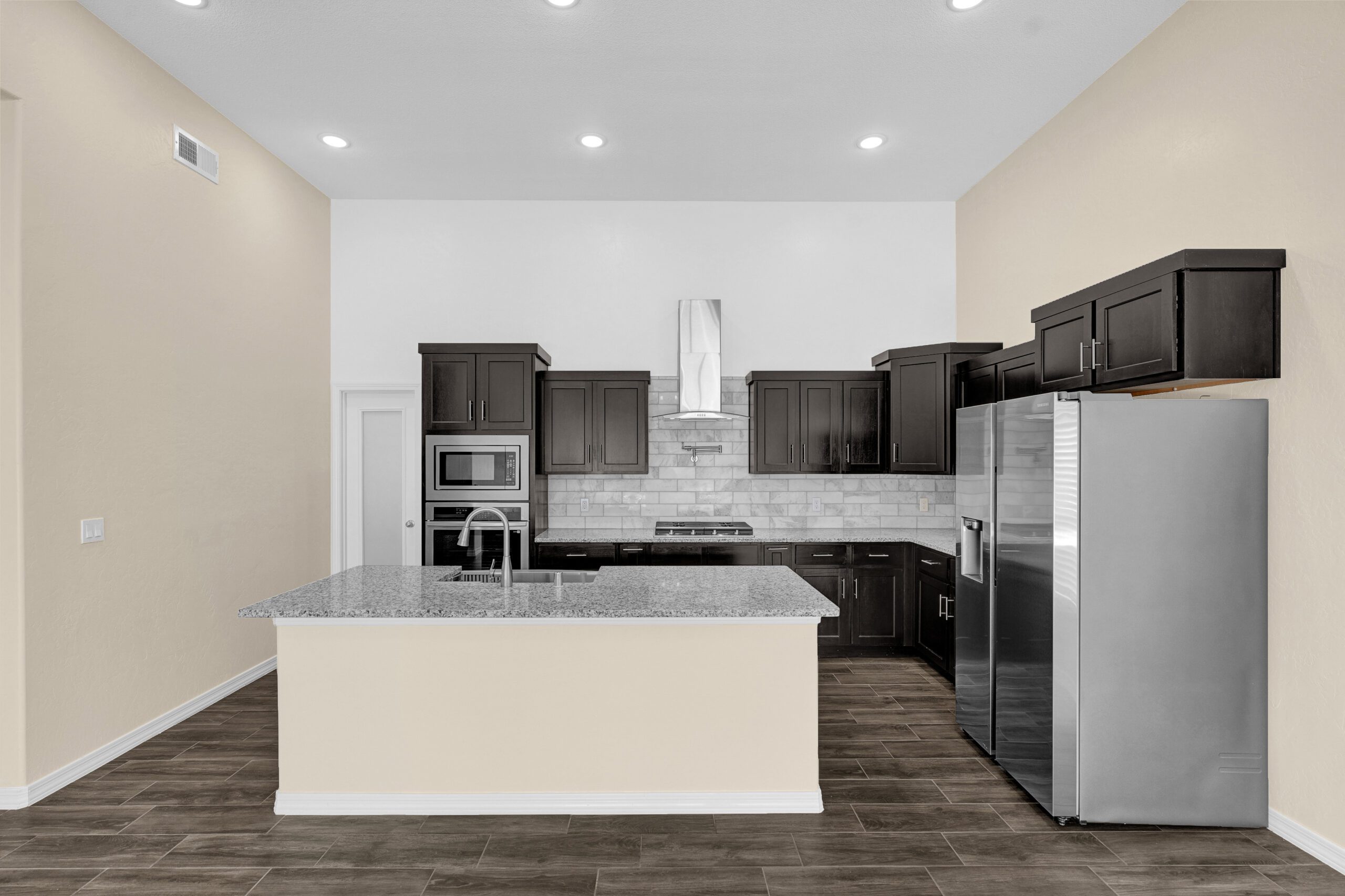 Front view of kitchen in The Lavender model by Zia Homes, featuring a central granite island, stainless steel appliances, and dark shaker-style cabinetry.
