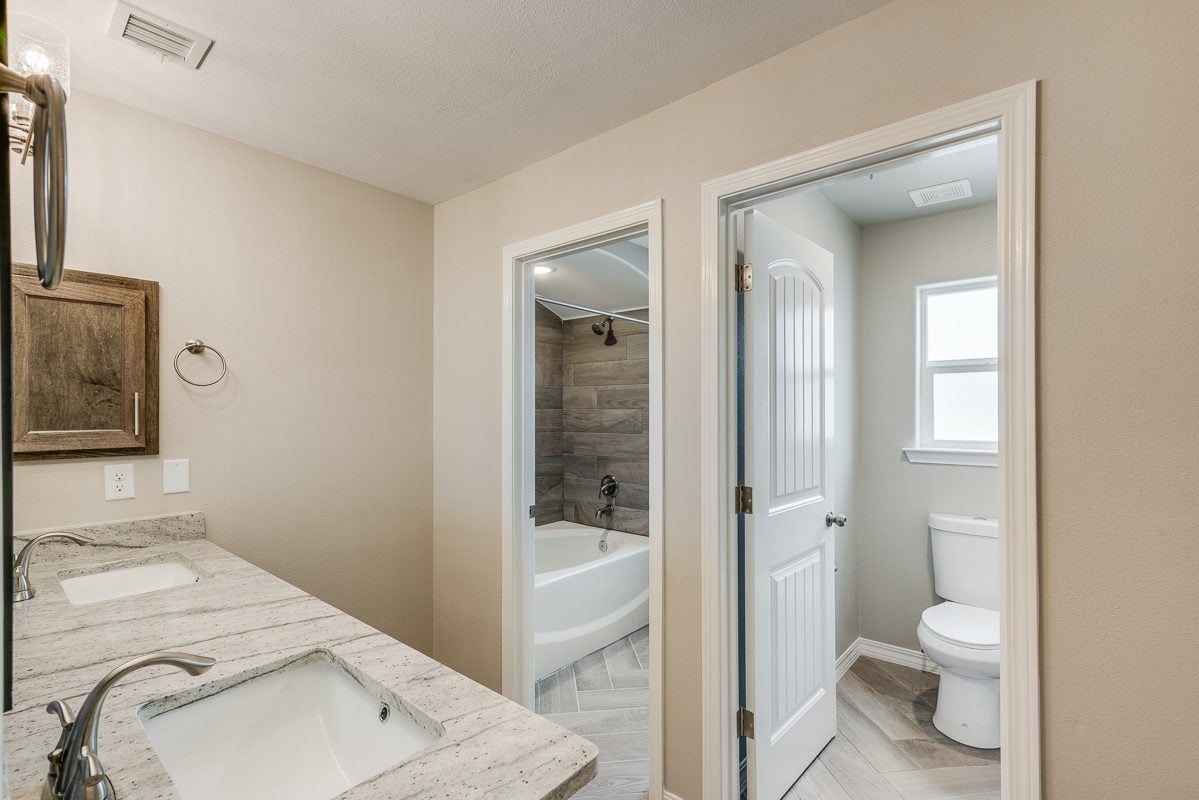 Secondary bathroom in The Chiffon featuring double sinks, private toilet room, and a bathtub with wood-look tile surround.