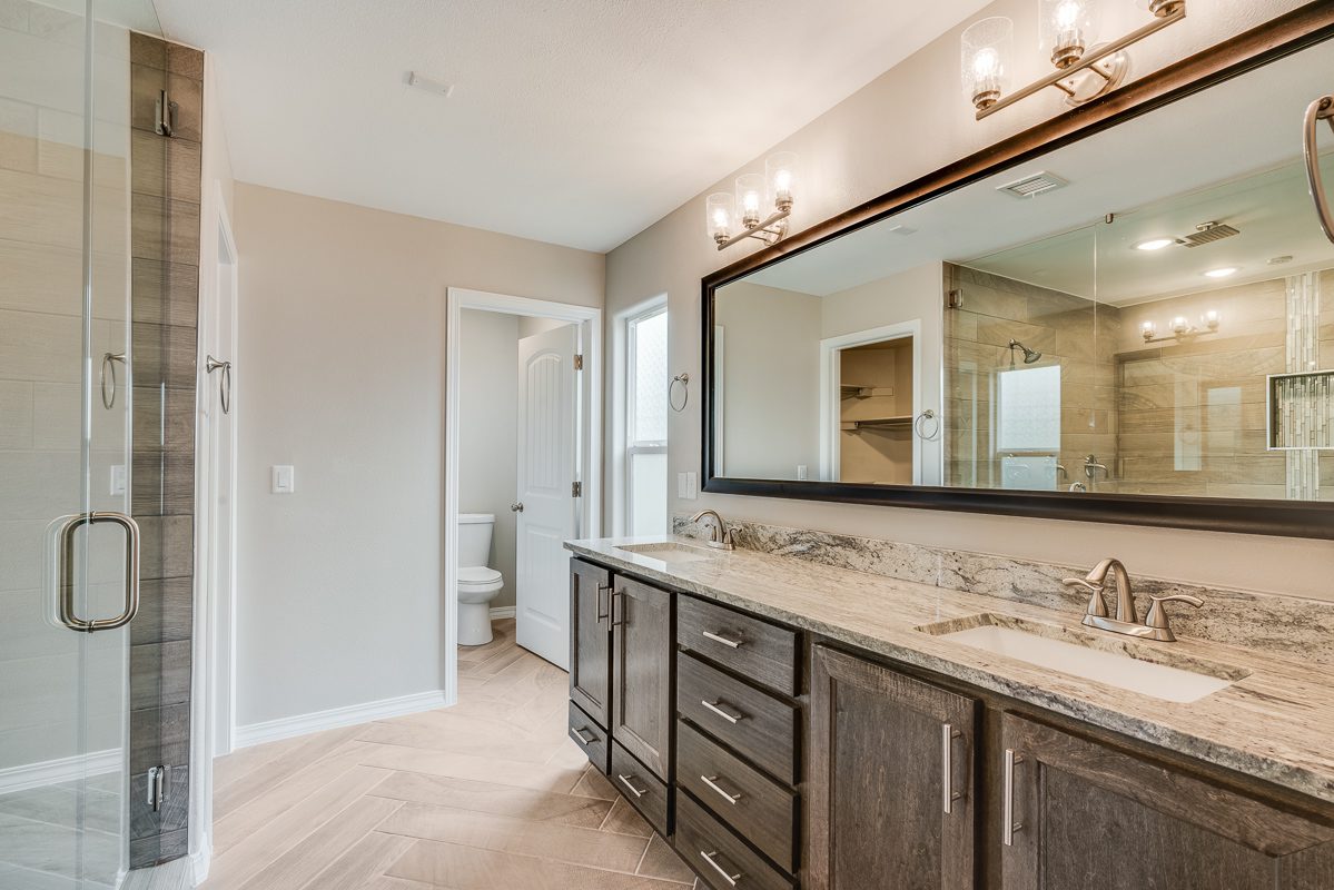 The Chiffon’s primary bathroom featuring a dual-sink vanity with granite countertops, dark wood cabinetry, large framed mirror, glass-enclosed shower, and natural wood-style tile flooring.