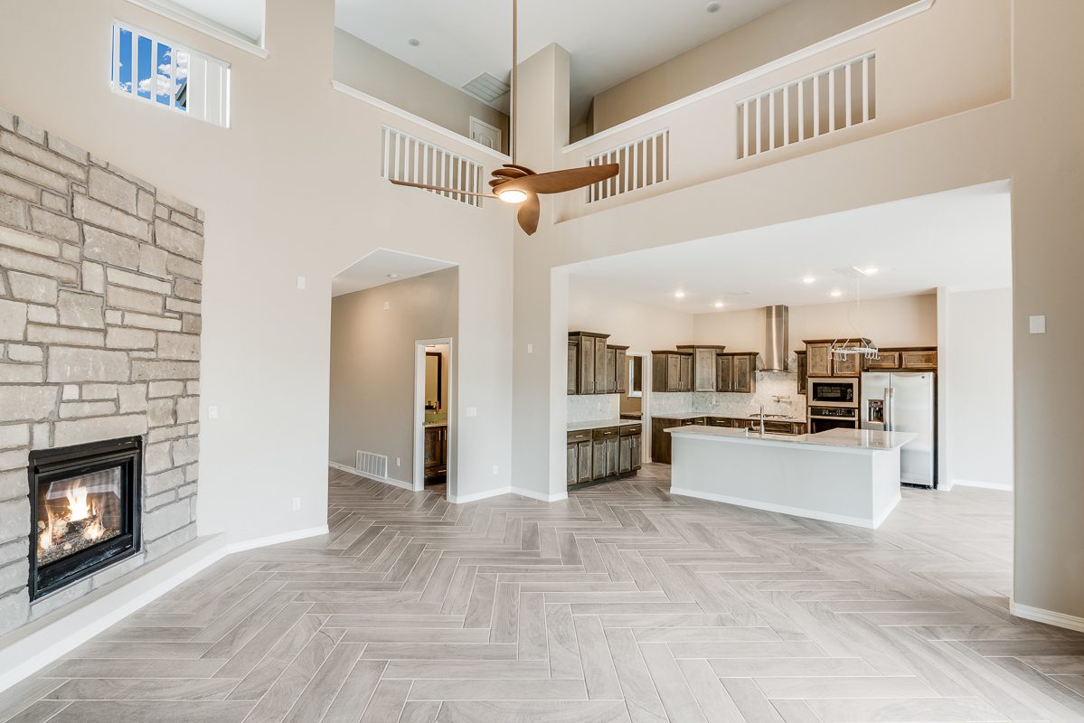 The Chiffon model open-concept living area featuring a grand two-story ceiling, modern fireplace, ceiling fan, and spacious kitchen with island and dark wood cabinetry.