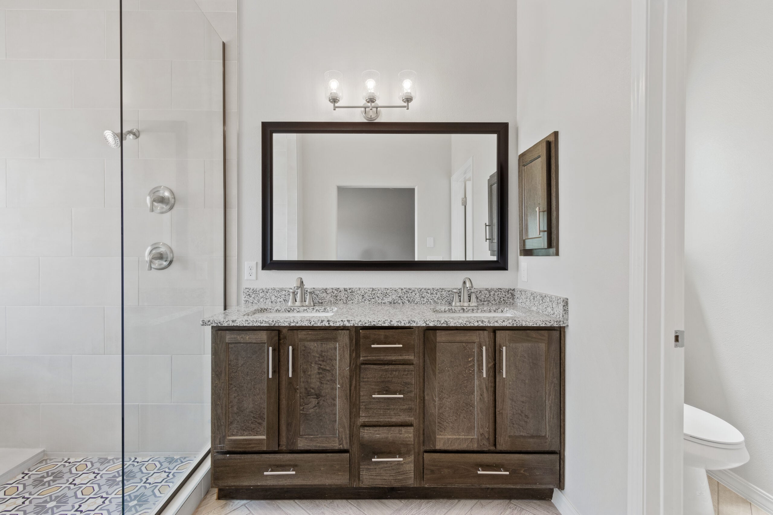 Modern Juniper bathroom with dual-sink granite vanity, framed mirror, and a walk-in glass shower featuring chrome fixtures.