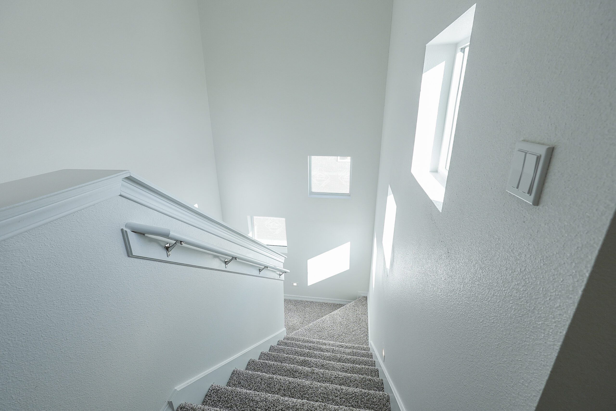 Modern carpeted staircase in The Playa home model by Zia Homes, illuminated by multiple rectangular windows with white walls.